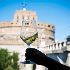 Un calice di vino bianco ondeggia con il logo "Vinea Domini" sullo sfondo di Castel Sant'Angelo a Roma.