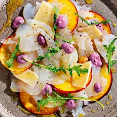 Delicato carpaccio di pesce, fettine di pesca, formaggio, rucola e sfere viola, un'interpretazione moderna dei frutti di mare crudi.