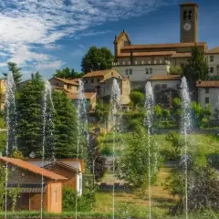Fontane zampillanti davanti a un borgo collinare con chiesa e campanile. Un'immagine che invita a scoprire cosa vedere a Fortunago.