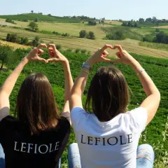 Due amiche formano cuori con le mani, con sullo sfondo le colline di Redavalle e i vigneti che daranno vita al vino.