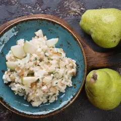Risotto pere e taleggio cremoso, guarnito con spicchi di pera fresca. Due pere verdi riposano su un tagliere di legno scuro.