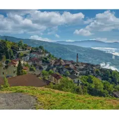 Un borgo di Pregola aggrappato alla collina, con case colorate e un campanile che svetta tra il verde. Nuvole basse accarezzano le montagne sullo sfondo.