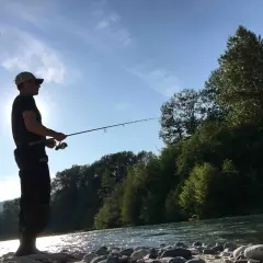 Uomo con cappellino pesca sul fiume Ticino, circondato da alberi e montagne. Un luogo ideale dove pescare sul Ticino.
