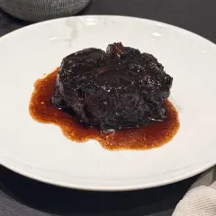 Un succulento brasato al buttafuoco, lucido e scuro, adagiato su un piatto bianco con un sugo denso e aromatico.