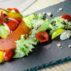 Un'insalata gourmet con terrina arancione, pomodorini colorati, ravanelli, lattuga e una salsa cremosa su una lavagna scura.
