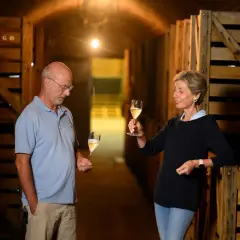 Coppia degusta vino in cantina, tra casse di legno. Il vino, affinato sur lie, brilla nei calici.