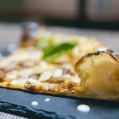 Una fetta di pizza al taglio con mandorle e basilico, servita su una lavagna scura con gocce di salsa.