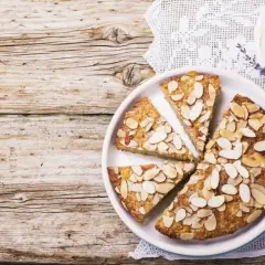 Fetta di torta di mandorle su un piatto bianco, guarnita con mandorle a lamelle. Sullo sfondo, un tovagliolo di pizzo e due uova di quaglia.