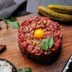 Tartare di carne cruda, con tuorlo d'uovo, cetriolini, pane tostato, sale grosso e prezzemolo su tagliere di legno.