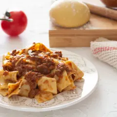 Piatto di pappardelle alla lepre, condite con un ricco ragù. Sullo sfondo, pomodori freschi, impasto e mattarello.