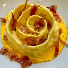 Pappardella ripiena a forma di rosa, adagiata su una crema gialla e guarnita con croccante prosciutto.