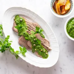 Lingua bollita affettata, generosamente condita con salsa verde e capperi. Accanto, una ciotola di salsa verde e altra guarnizione.