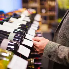 Un uomo in giacca sceglie una bottiglia di vino, forse un pregiato vino muffato, tra una fila di etichette bianche.