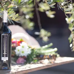 Bottiglia di vino Grechetto Colli Martani, con fiori e occhiali da sole su un tavolo di legno, sotto i rami di un ulivo.