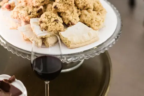 A glass of red wine sits next to a cake stand filled with cookies and pastries.