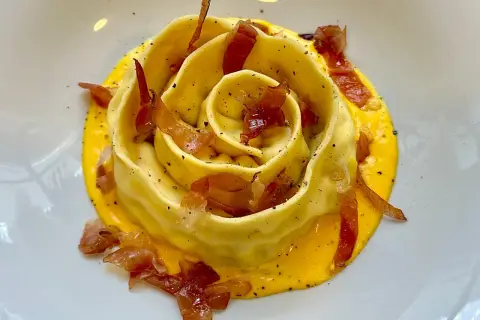 Pappardella ripiena a forma di rosa, adagiata su una crema gialla e guarnita con croccante prosciutto.