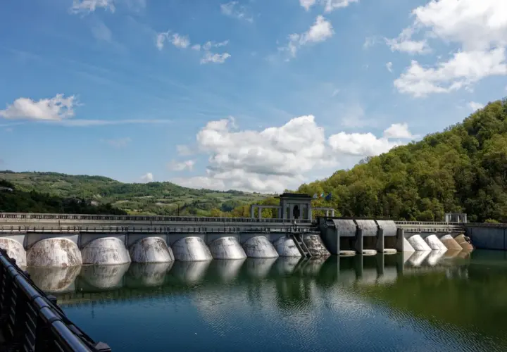 Diga imponente con colline verdi sullo sfondo, ideale per il trekking a Pavia. L'acqua riflette il cielo azzurro e le nuvole bianche.