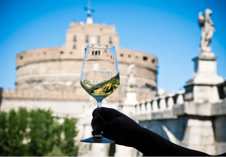 Un calice di vino bianco ondeggia con il logo "Vinea Domini" sullo sfondo di Castel Sant'Angelo a Roma.