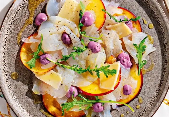 Delicato carpaccio di pesce, fettine di pesca, formaggio, rucola e sfere viola, un'interpretazione moderna dei frutti di mare crudi.