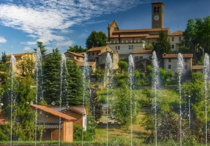 Fontane zampillanti davanti a un borgo collinare con chiesa e campanile. Un'immagine che invita a scoprire cosa vedere a Fortunago.