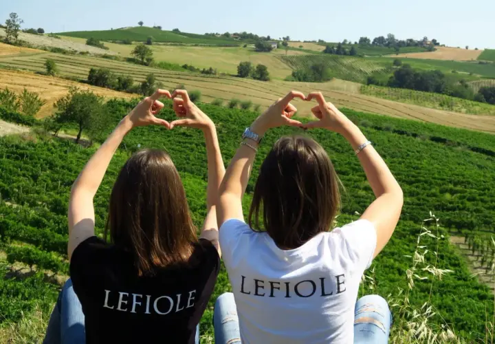 Due amiche formano cuori con le mani, con sullo sfondo le colline di Redavalle e i vigneti che daranno vita al vino.