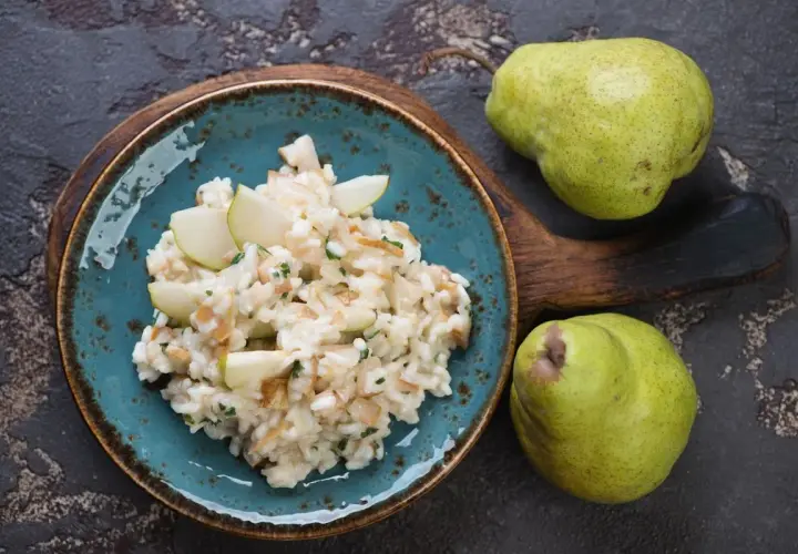 Risotto pere e taleggio cremoso, guarnito con spicchi di pera fresca. Due pere verdi riposano su un tagliere di legno scuro.