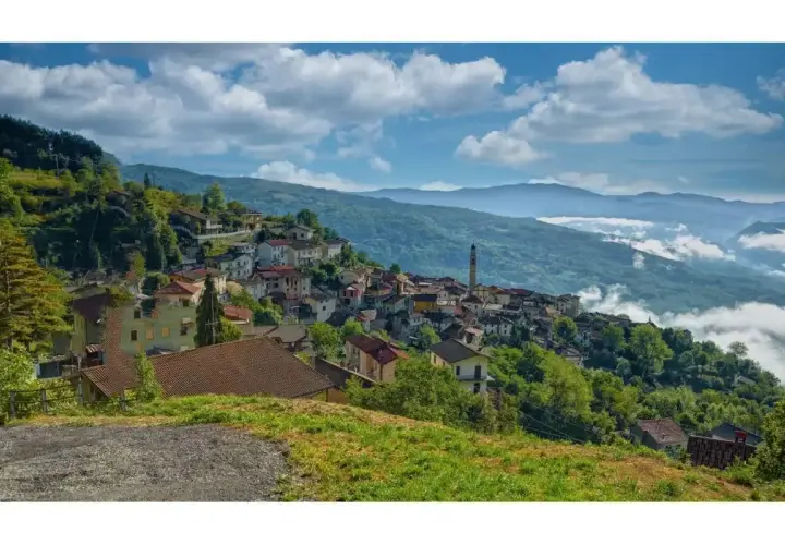 Un borgo di Pregola aggrappato alla collina, con case colorate e un campanile che svetta tra il verde. Nuvole basse accarezzano le montagne sullo sfondo.