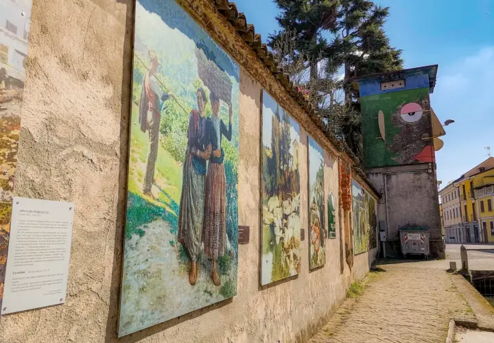 Opere d'arte lungo un muro a Gravellona Lomellina, con dipinti che raffigurano scene di vita rurale. Un'installazione artistica colorata completa il panorama.