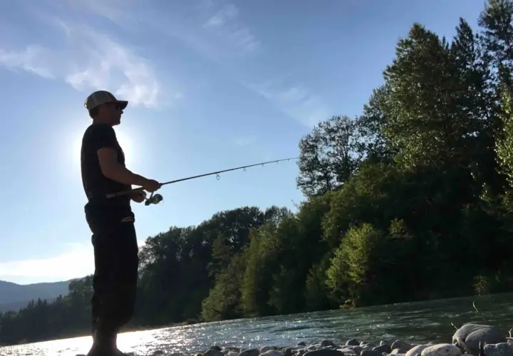 Uomo con cappellino pesca sul fiume Ticino, circondato da alberi e montagne. Un luogo ideale dove pescare sul Ticino.