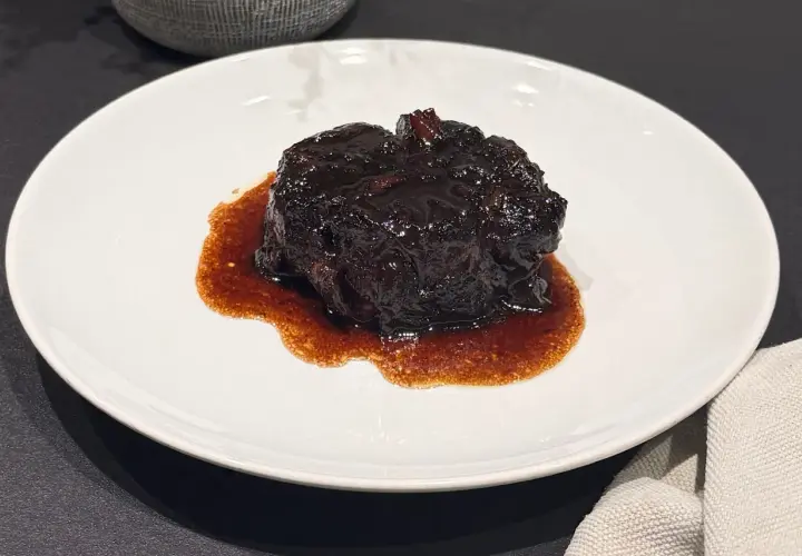 Un succulento brasato al buttafuoco, lucido e scuro, adagiato su un piatto bianco con un sugo denso e aromatico.