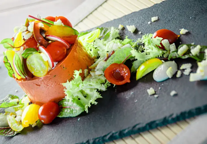 Un'insalata gourmet con terrina arancione, pomodorini colorati, ravanelli, lattuga e una salsa cremosa su una lavagna scura.