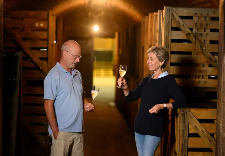 Coppia degusta vino in cantina, tra casse di legno. Il vino, affinato sur lie, brilla nei calici.