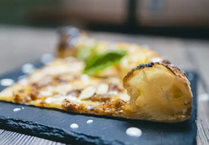 Una fetta di pizza al taglio con mandorle e basilico, servita su una lavagna scura con gocce di salsa.