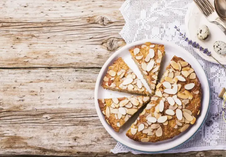 Fetta di torta di mandorle su un piatto bianco, guarnita con mandorle a lamelle. Sullo sfondo, un tovagliolo di pizzo e due uova di quaglia.