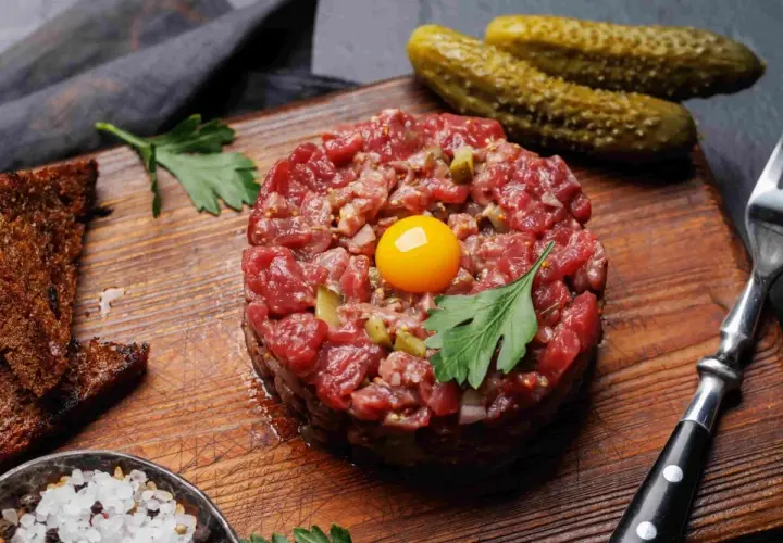Tartare di carne cruda, con tuorlo d'uovo, cetriolini, pane tostato, sale grosso e prezzemolo su tagliere di legno.