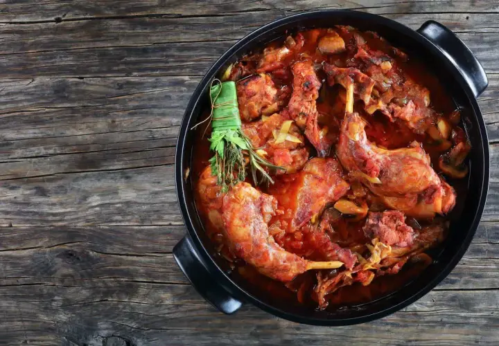 Un saporito coniglio al vino rosso, cotto lentamente con erbe aromatiche, in una casseruola nera su un tavolo di legno rustico.