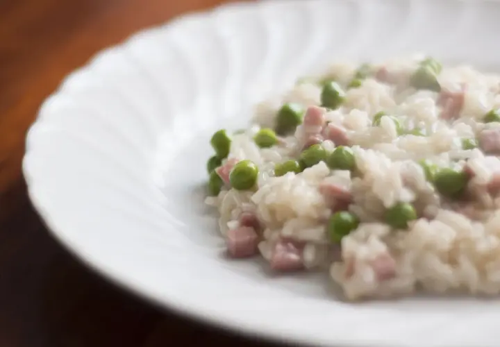 Un cremoso riso con piselli e cubetti di prosciutto, servito su un piatto bianco con un bordo ondulato.