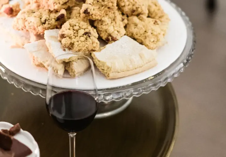 A glass of red wine sits next to a cake stand filled with cookies and pastries.