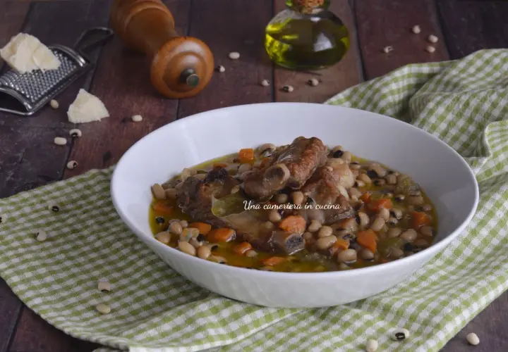 Una ciotola di zuppa dei morti con fagioli, costine di maiale e carote, servita su una tovaglia a quadri verdi.