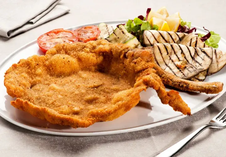Cotoletta alla milanese dorata e croccante, servita con pomodori grigliati, melanzane e un'insalata fresca.