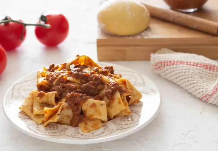 Piatto di pappardelle alla lepre, condite con un ricco ragù. Sullo sfondo, pomodori freschi, impasto e mattarello.
