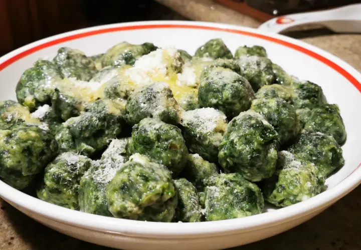 Malfatti ricetta originale: gnocchetti verdi fatti in casa, conditi con burro fuso e parmigiano grattugiato, serviti in una ciotola bianca con bordo rosso.