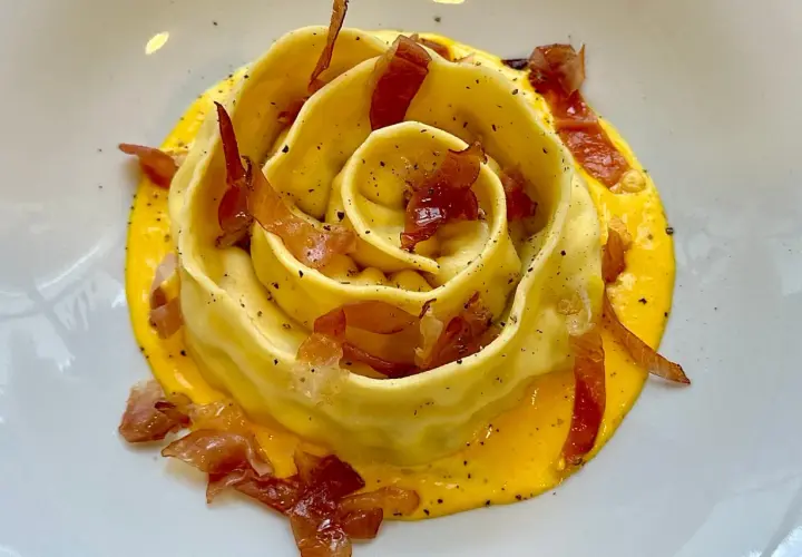 Pappardella ripiena a forma di rosa, adagiata su una crema gialla e guarnita con croccante prosciutto.
