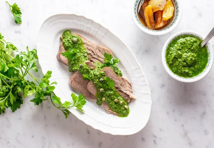 Lingua bollita affettata, generosamente condita con salsa verde e capperi. Accanto, una ciotola di salsa verde e altra guarnizione.