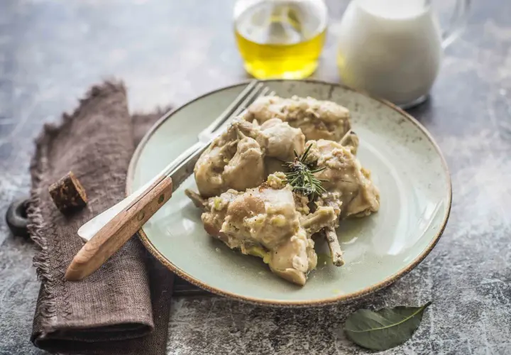 Piatto di coniglio al latte, tenero e saporito, guarnito con rosmarino. Accanto, olio e latte in caraffa.