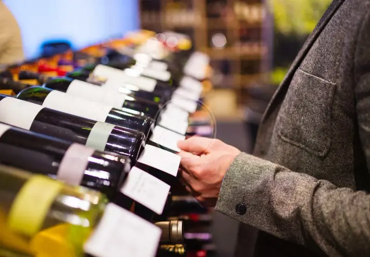 Un uomo in giacca sceglie una bottiglia di vino, forse un pregiato vino muffato, tra una fila di etichette bianche.
