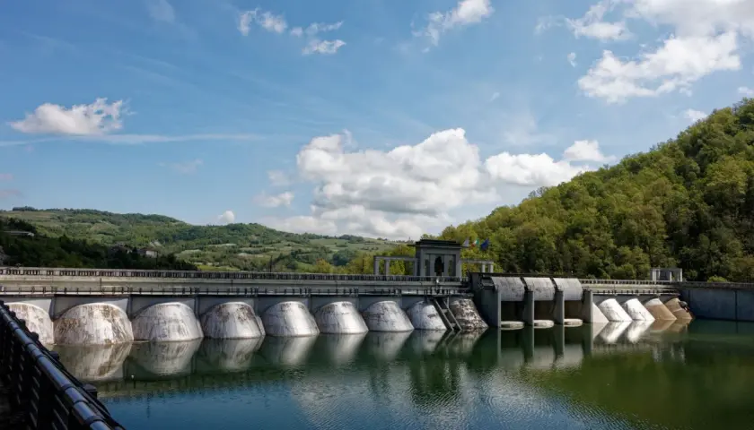 Diga imponente con colline verdi sullo sfondo, ideale per il trekking a Pavia. L'acqua riflette il cielo azzurro e le nuvole bianche.