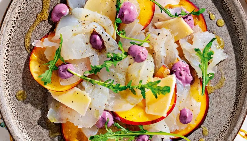 Delicato carpaccio di pesce, fettine di pesca, formaggio, rucola e sfere viola, un'interpretazione moderna dei frutti di mare crudi.