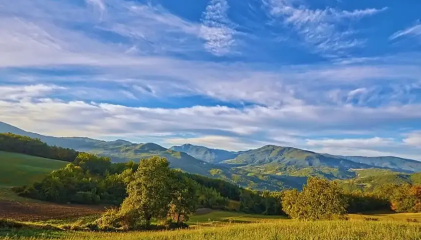 Valli della Staffora sotto un cielo azzurro con nuvole bianche. Colline verdi e alberi in primo piano.