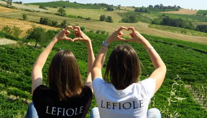 Due amiche formano cuori con le mani, con sullo sfondo le colline di Redavalle e i vigneti che daranno vita al vino.